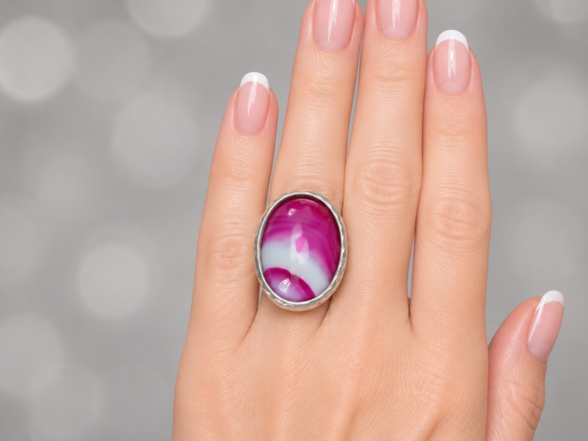 A close-up of a hand wearing a silver ring with an oval cabochon featuring a marbled pink, purple, and white pattern, set against a soft gray bokeh background.