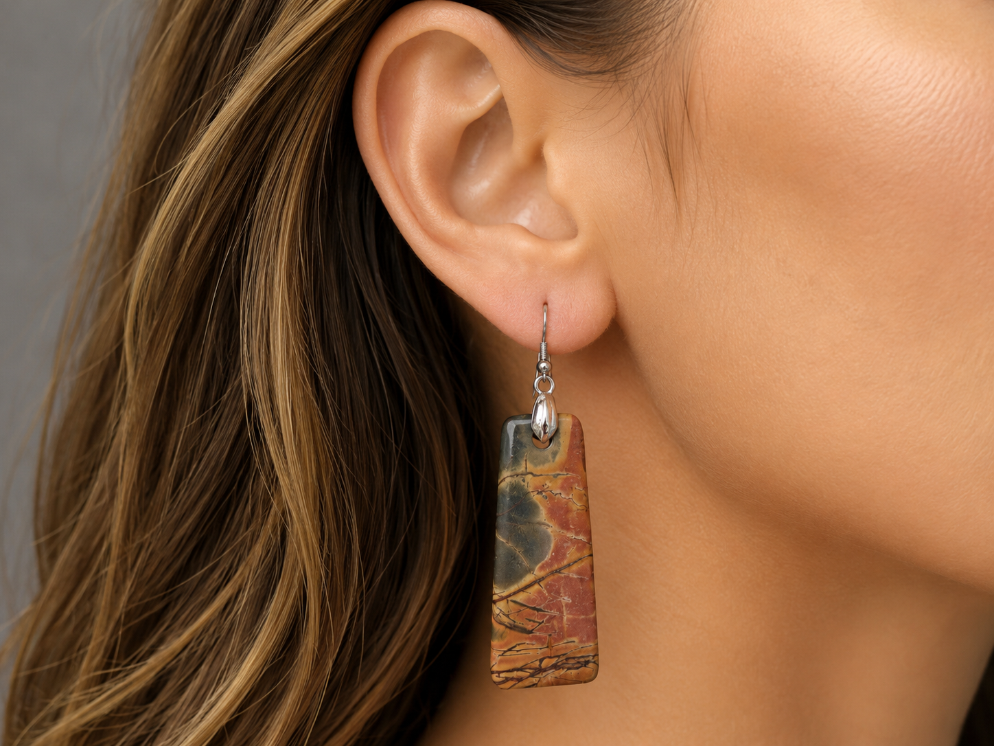 A close-up of a woman's ear wearing a rectangular drop earring made from a marbled stone with brown, gold, and gray tones, suspended from a silver fishhook. Main image