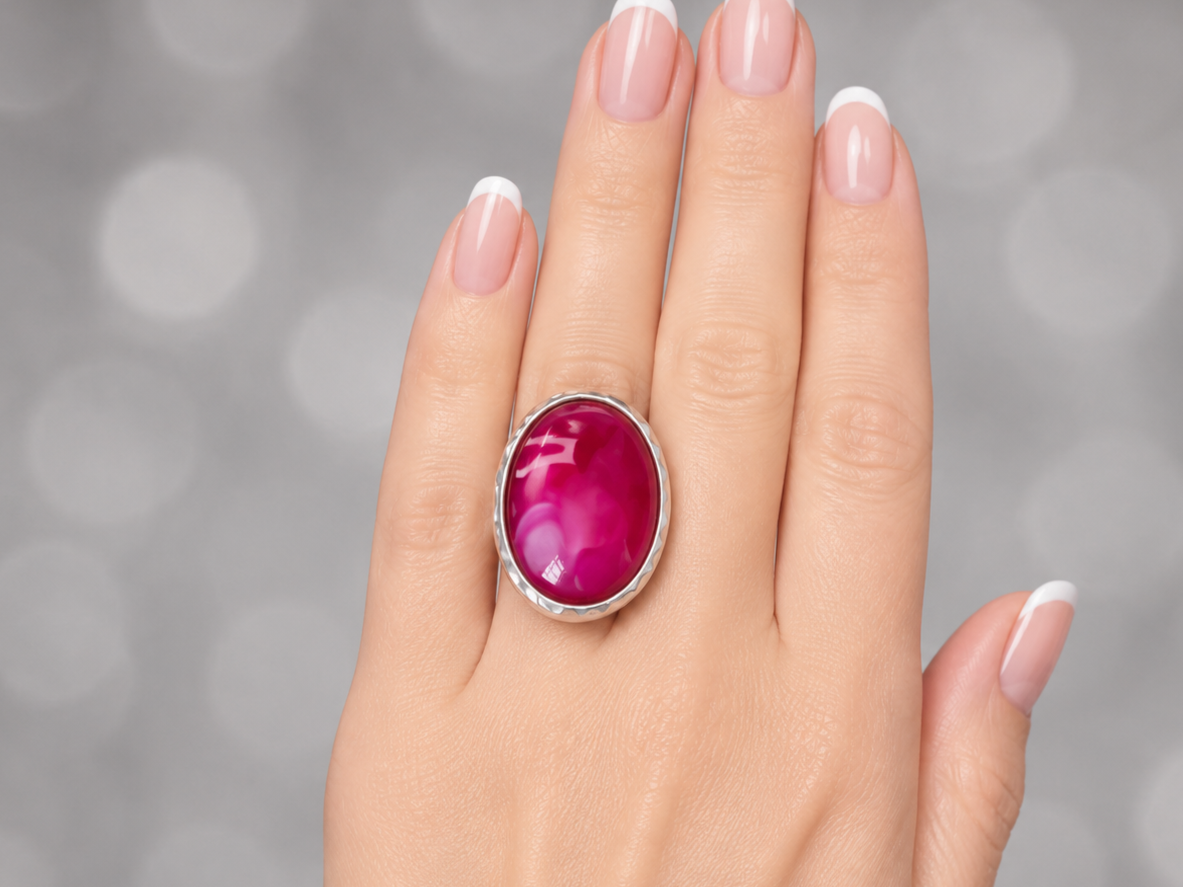 A close-up of a hand with a French manicure wearing a silver ring with a large, oval, magenta-pink cabochon gemstone, set against a soft, out-of-focus gray bokeh background.