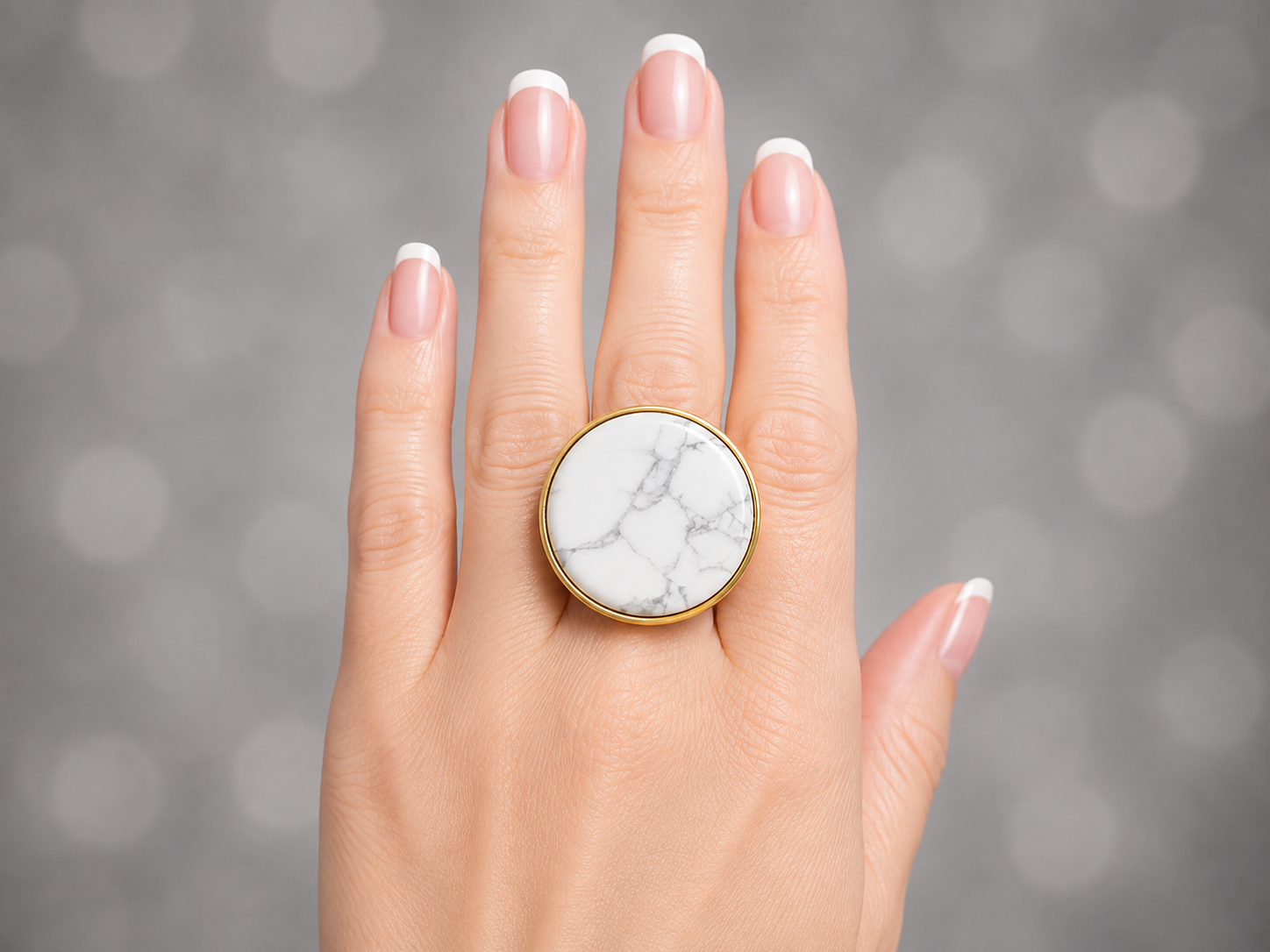A close-up of a hand with a French manicure wearing a large round ring with a white and gray marble inlay and a gold-tone bezel, set against a soft gray bokeh background.