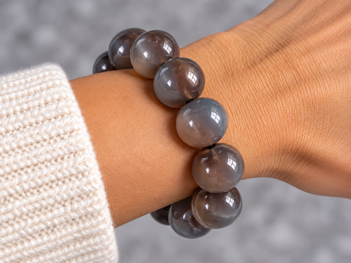 A close-up of a person's wrist wearing a beaded bracelet made of smooth, polished oval beads in a smoky gray and translucent brown color, paired with a cream-colored knit sweater cuff.