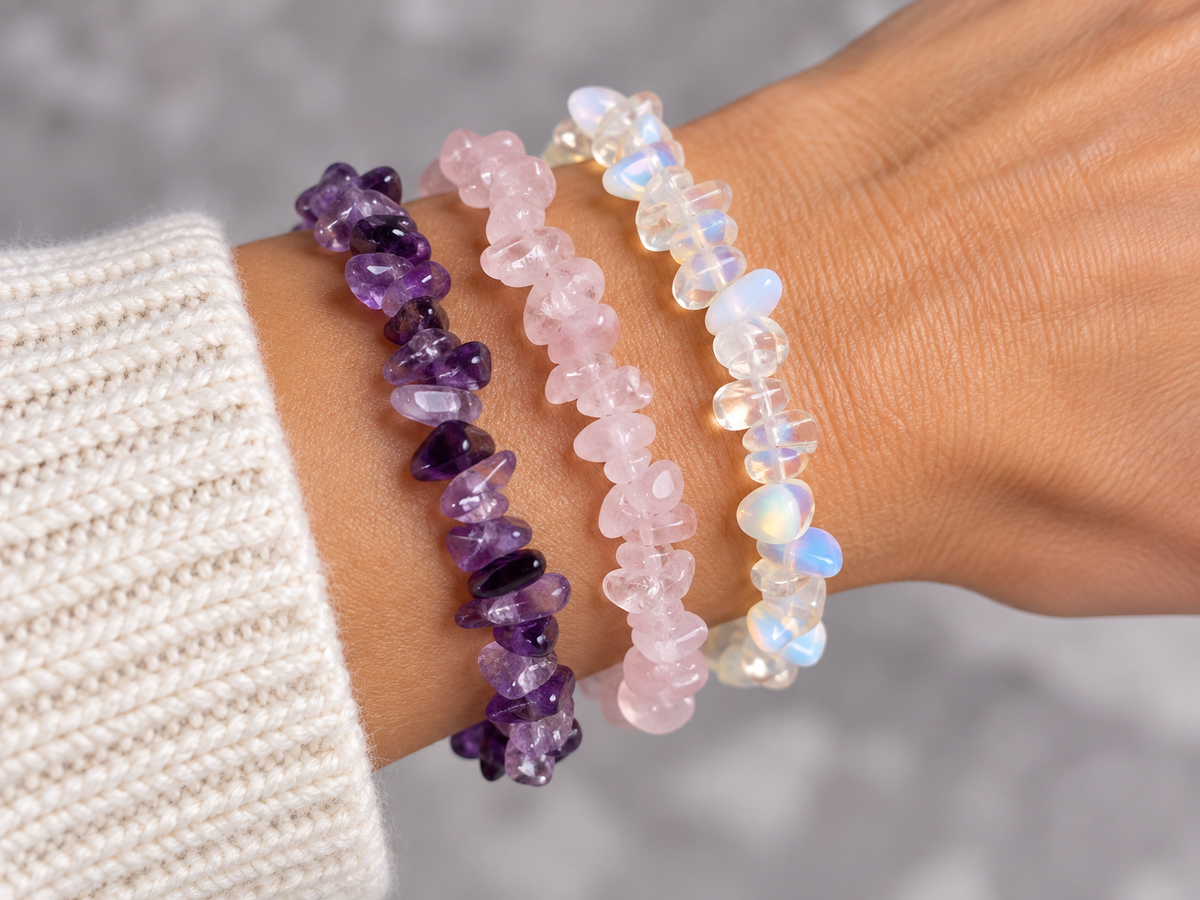 Three beaded stretch bracelets worn on a wrist: a purple amethyst chip bracelet, a light pink rose quartz chip bracelet, and a white iridescent moonstone chip bracelet. A cream knit sleeve is visible on the left against a soft gray background.