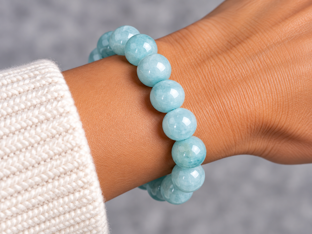 A close-up of a person's wrist wearing a double-strand beaded bracelet made of light blue faceted gemstone beads, paired with a white knit sleeve, set against a soft gray background.
