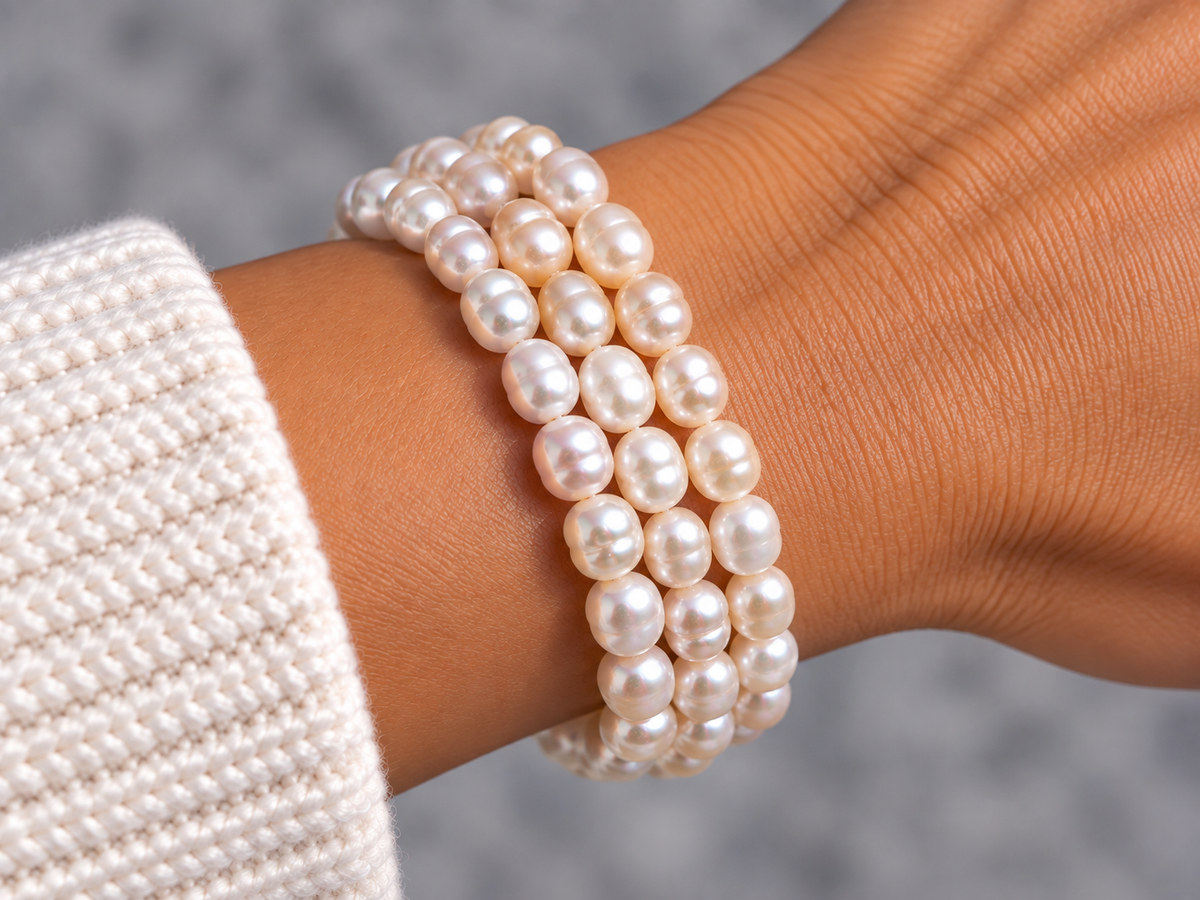 A close-up of a person's wrist wearing a three-strand pearl bracelet, with a cream knit sleeve visible on the left against a soft gray background.