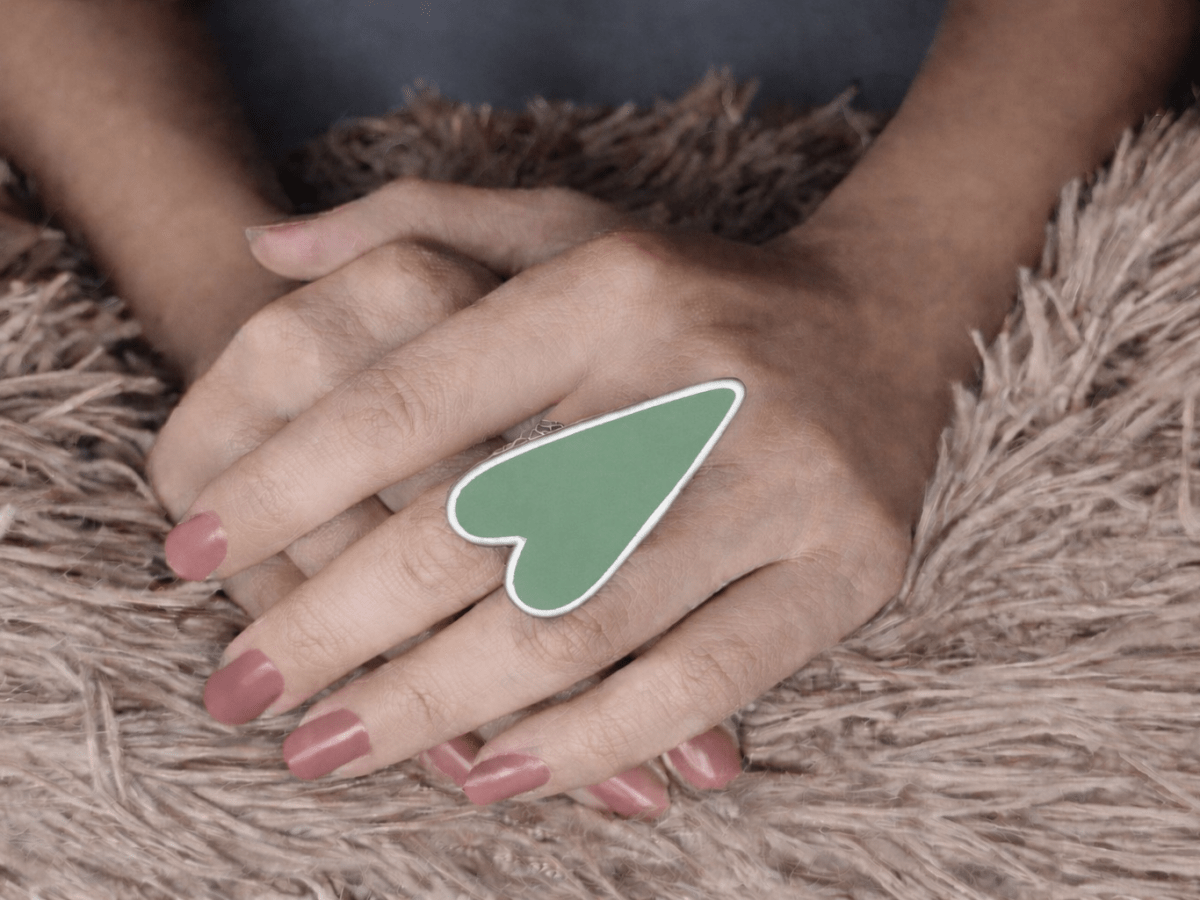 A models hand wearing a large sage green heart shaped ring